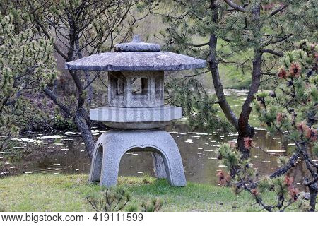 Japanese Garden At Spring, Oriental Culture. Old Yukimi-toro Stone Lantern On Island In Pond Surroun