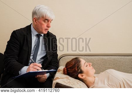 Caucasian Woman Lies On A Couch At A Reception With A Psychotherapist. An Elderly Man Works As A Psy