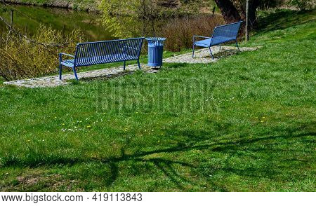 Park Benches In Light Blue Made Of Metal Strips Similar To A Lattice. Trash Can And Beautiful Lawn W