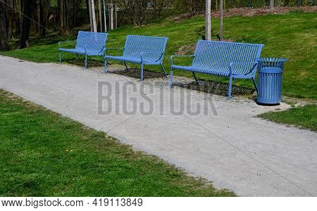 Park Benches In Light Blue Made Of Metal Strips Similar To A Lattice. Trash Can And Beautiful Lawn W