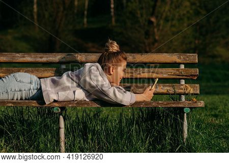 A Blonde Girl Looks On The Phone On The Park Bench