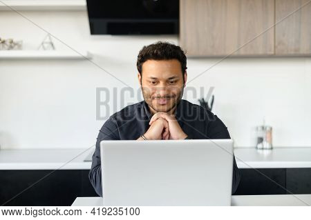 Front View Of Positive Hindoo Man In Smart Casual Shirt Using Laptop Sitting At The Desk In The Kitc