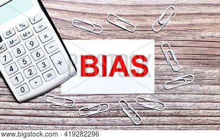 On A Wooden Background, A White Calculator, White Paper Clips And A White Card With The Text Bias. V