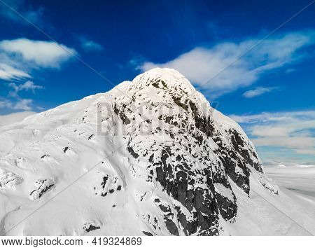 Panoramic View Of A Snow Capped Mountain Peak With Clouds And Blue Skies.