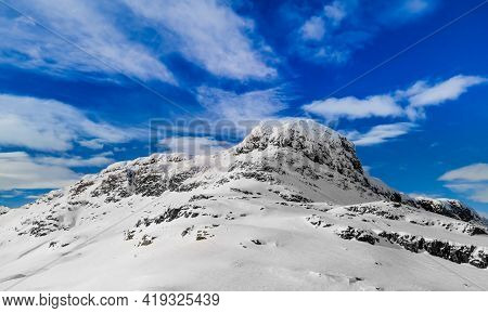Panoramic View Of A Snow Capped Mountain Peak With Clouds And Blue Skies.