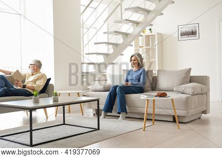 Older Senior Family Couple Spending Time At Home In Modern Living Room. Middle Aged Husband Reading 