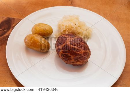 Saumaise, A Pork Caul Filled With Cured Meat, With Sauerkraut And New Potatos On A White Plate, A Tr