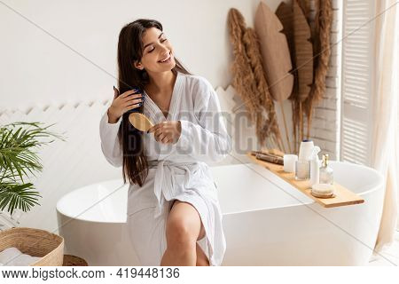 Brunette Female Brushing Hair With Hairbrush Sitting On Bathtub Indoor