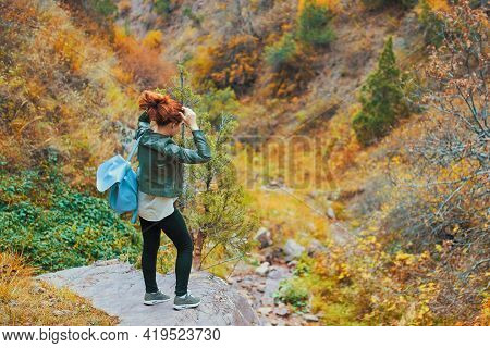 Female Tourist With A Backpack Admires The Mountain Scenery. A Red-haired Woman Ties Her Hair On The