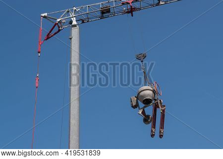 A Round Grey Concrete Mixer And A Red Movable Carriage Trolley Are Suspended From The Boom Hook Of A