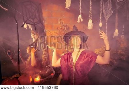 Blonde Witch In Red Dress And Black Hat With Candle And Mirror In Halloween Decoration Indoors.