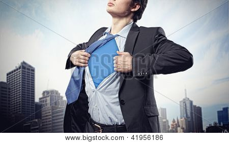 Image of young businessman showing superhero suit underneath his shirt standing against city background