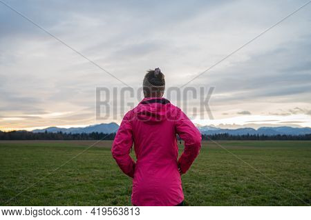 View From Behind Of A Woman In Bright Pink Jacket Standing In Beautiful Meadow Under An Evening Sky 
