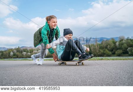 Laughing Kids Have Fun Using A Skateboard. Brother Pushing His Sister Sitting On A Skateboard And Ma