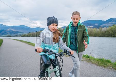 Laughing Kids Have Fun Using A Skateboard. Brother Pushing His Sister Sitting On A Skateboard And Ma