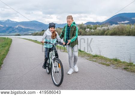 Laughing Kids Have Fun Using A Skateboard. Brother Pushing His Sister Sitting On A Skateboard And Ma