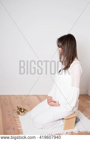 Beautiful Adult Woman Dressed In White During A Meditation Sesion. Wooden Bench And Cupper Singing B