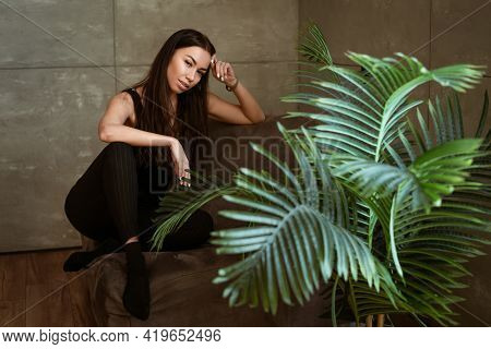 Beautiful Young Woman Closeup Portrait Through Green Leaves Of A Flower On A Gray Wall Background