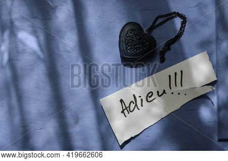 Handwritten Word Adieu In A White Piece Of Paper And A Wooden Amulet In The Shape Of A Heart. The Wo