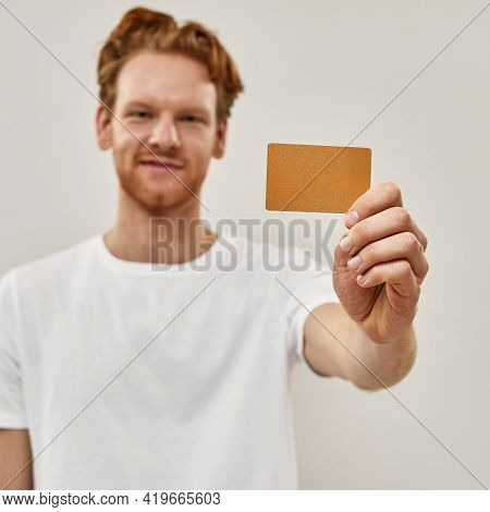 Young Redhead Man Holds Credit Card In Front Of Him, Focus Cought On The Credit Card , Man Smiles An