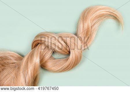 Blonde Hair Lock Tied In Knot. Strand Of Honey Blonde Hair On Mint Background, Top View. Hairdresser