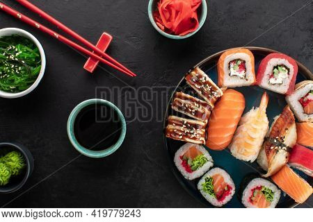 Assorted sushi roll set served on a blue round plate, seaweed salad, soy sauce and ginger in a bowl on the side. Asian traditional food on black background, top view