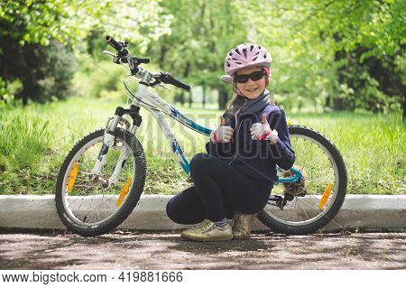 Konstantinovka, Ukraine - May 8, 2021: Little Girl Near The Bike Holds Thumbs Up. Warm Sunny Summer 