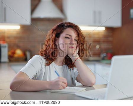 A Beautiful Young Redheaded Woman Sits In The Kitchen At The Table With Her Laptop.