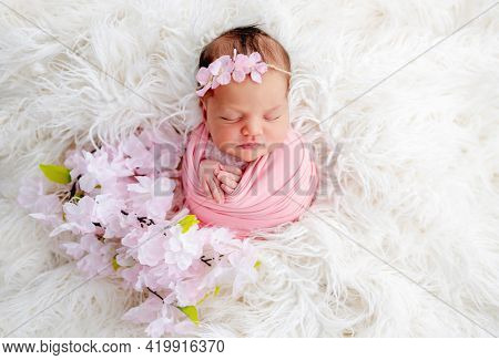 Swaddled beautiful newborn baby girl with flower wreath on her head sleeping in white fur. Cute female infant child photoshoot in studio. Adorable kid napping