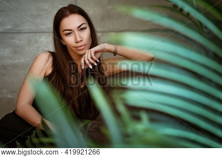 Beautiful Young Woman Closeup Portrait Through Green Leaves Of A Flower On A Gray Wall Background
