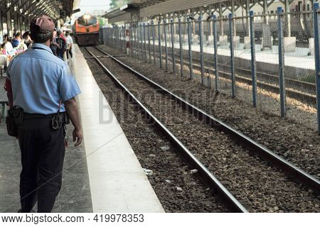 Rear View Of Security Guard In The Uniform Works At The Platform Of Train Station While The Train Is