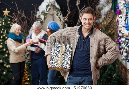 Portrait of happy young woman holding Christmas present with family standing in background at store