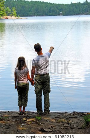Father and daughter standing on the lake shore and looking at calm water, father pointing at something on the other shore