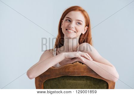 Smiling redhead woman sitting on the chair and looking away isolated on a white background