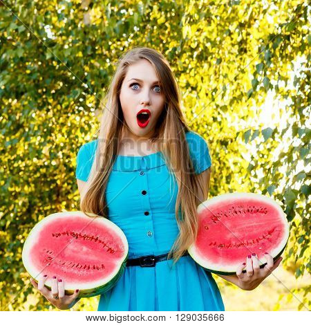 Beautiful blonde girl in a blue dress with long hair holding two halves of a sliced watermelon