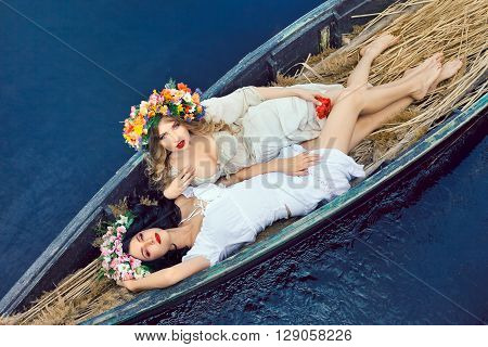 Two young women on boat at sunset. The girls has a colorfull make-up and flower wreath on they head, relaxing and sailing on the river. Beautiful sexy slim body and cute face.  Concept of female beauty, rest on the nature, and wate