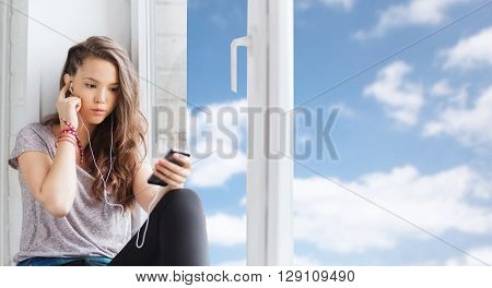 people, technology and teens concept - sad pretty teenage girl sitting on windowsill with smartphone and earphones listening to music over blue sky and clouds background