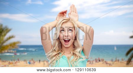 fun, expressions, summer holidays, travel and people concept - happy smiling young woman making bunny ears over exotic tropical beach with palm trees and sea background