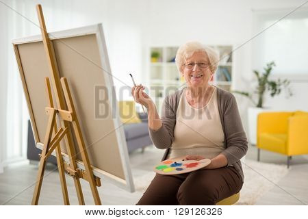 Elderly woman painting on a canvas seated on a chair at home