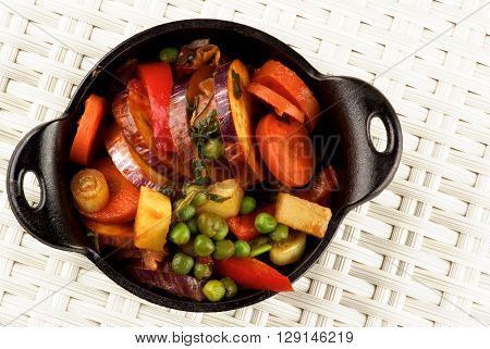 Delicious Homemade Colorful Vegetables Ragout with Eggplant Carrots Potatoes Leek Red Bell Pepper and Green Pea in Black Iron Stewpot closeup on Wicker background. Top View