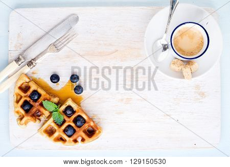 Espresso coffee cup and soft belgian waffles with fresh blueberries and marple syrup on white painted wooden board over light blue background. Top view, copy space, horizontal