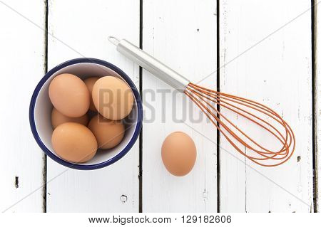 Eggs in a ceramic bowl with a wisk on a rustic white painted wood table top shot from above