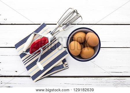 Vintage egg whisk with eggs in enamel bowl on a rustic retro white wooden background