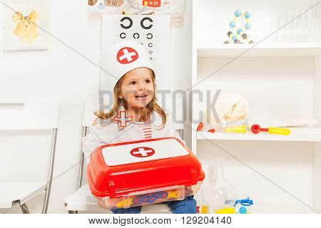 Cute little girl dressed like a doctor holding toy first-aid chest, sitting at the medical room