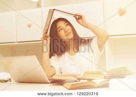Toned image. Happy Asian student holding exercise-book above her head while sitting in library and prepaing for lessons, classes or exams.