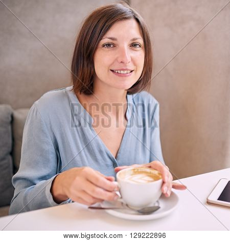 Caucasian woman with brown hair and a nice grey top smiling while looking at the camera as she has both hands on her fresh morning cup of coffee, her tablet just visible on the table infront of her