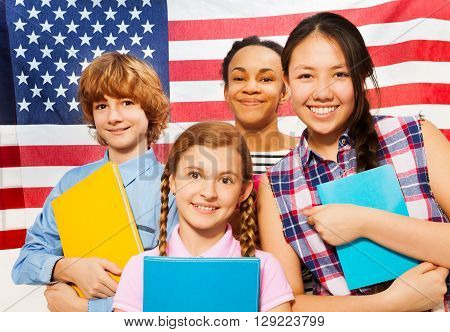 Close-up picture of four smiling multiethnic teenage students standing with textbooks against American flag