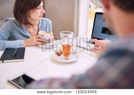 Tight shot of woman looking at laptop over mans shoulder in a coffee shop with phone and notebook lying on the table infront of them.