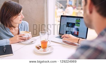 Tight shot of woman looking at laptop over mans shoulder in a coffee shop with phone and notebook lying on the table infront of them.