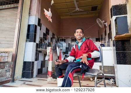 DARAW, EGYPT - FEBRUARY 6, 2016: Young local butcher sitting on the chair holding knife in front of butcher shop.